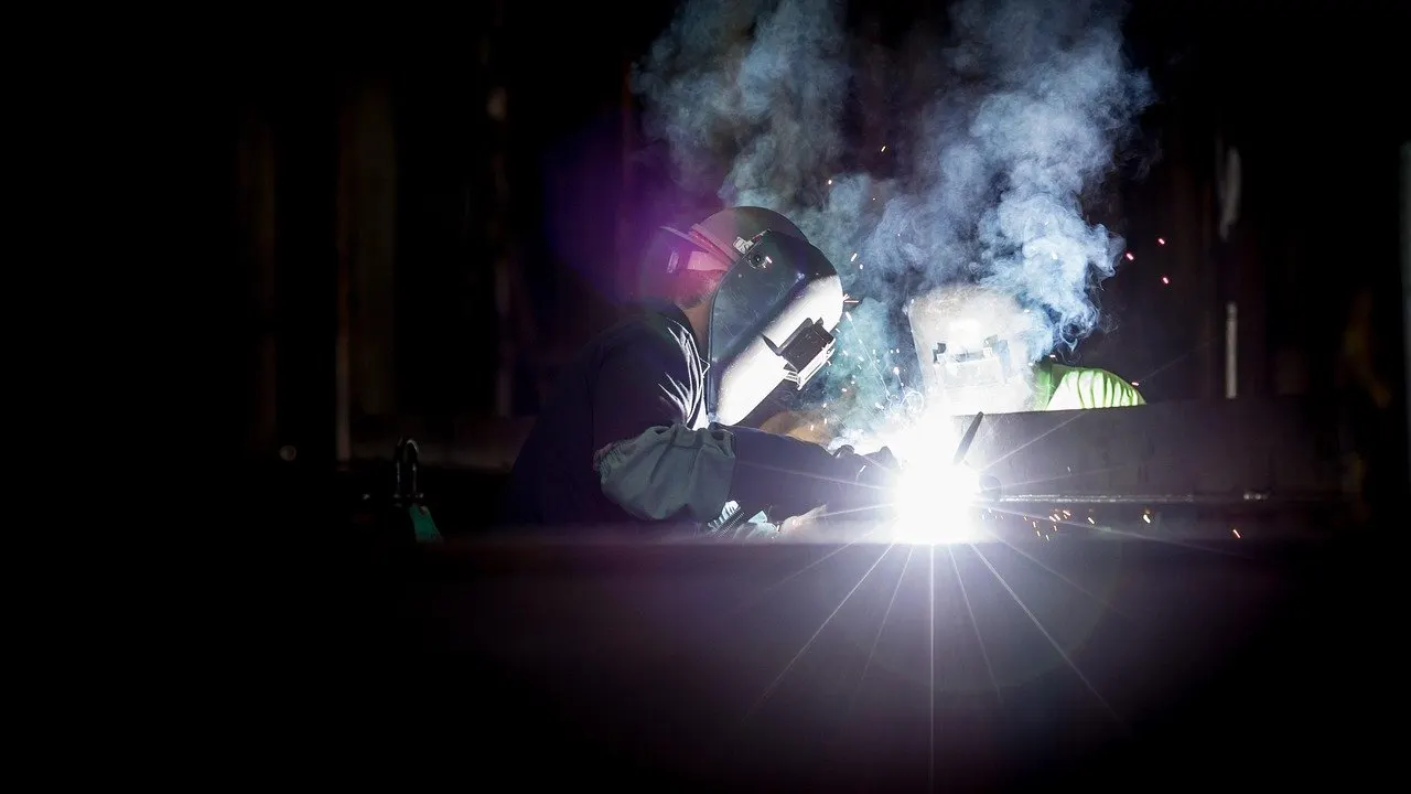 Schweißer mit Schutzhelm arbeitet nachts an Metallteil, helle Lichtblitze und Funkenflug.