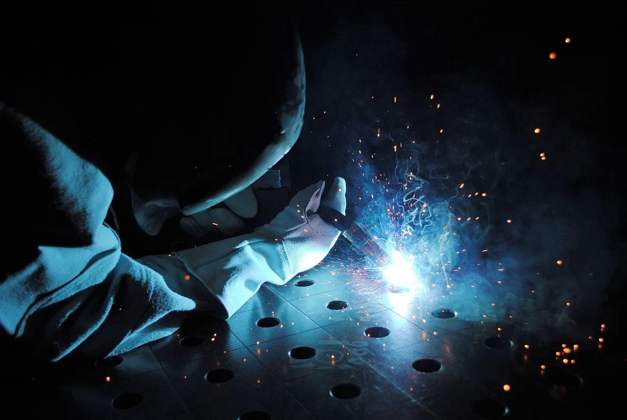 Schweißer mit Handschuh arbeitet im Dunkeln an einer gelochten Metallplatte, blauer Lichtbogen und Funken sind sichtbar.