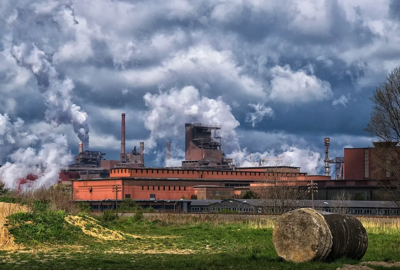 Industrieanlage mit Schornsteinen und Dampfwolken unter bewölktem Himmel, davor Wiese und Heuballen.