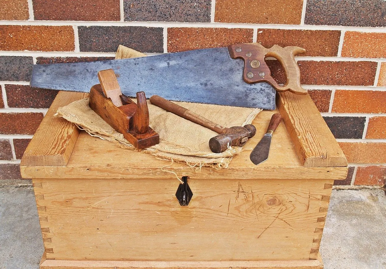 Alte Holzhandsäge, Hobel und Hammer liegen auf einer Holztruhe vor einer Ziegelwand.