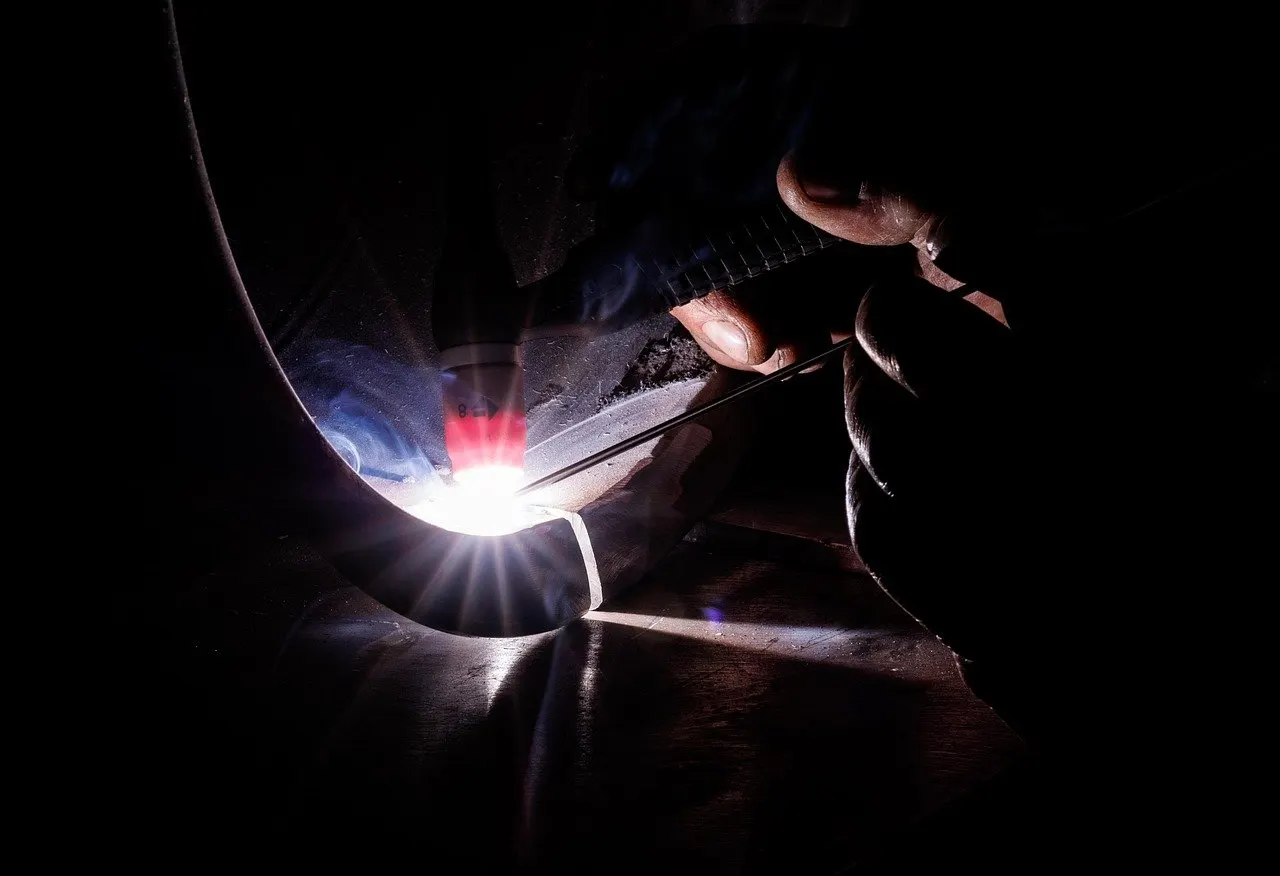 Schweißer arbeitet mit leuchtendem Lichtbogen an einem gebogenen Metallrohr in dunkler Umgebung.
